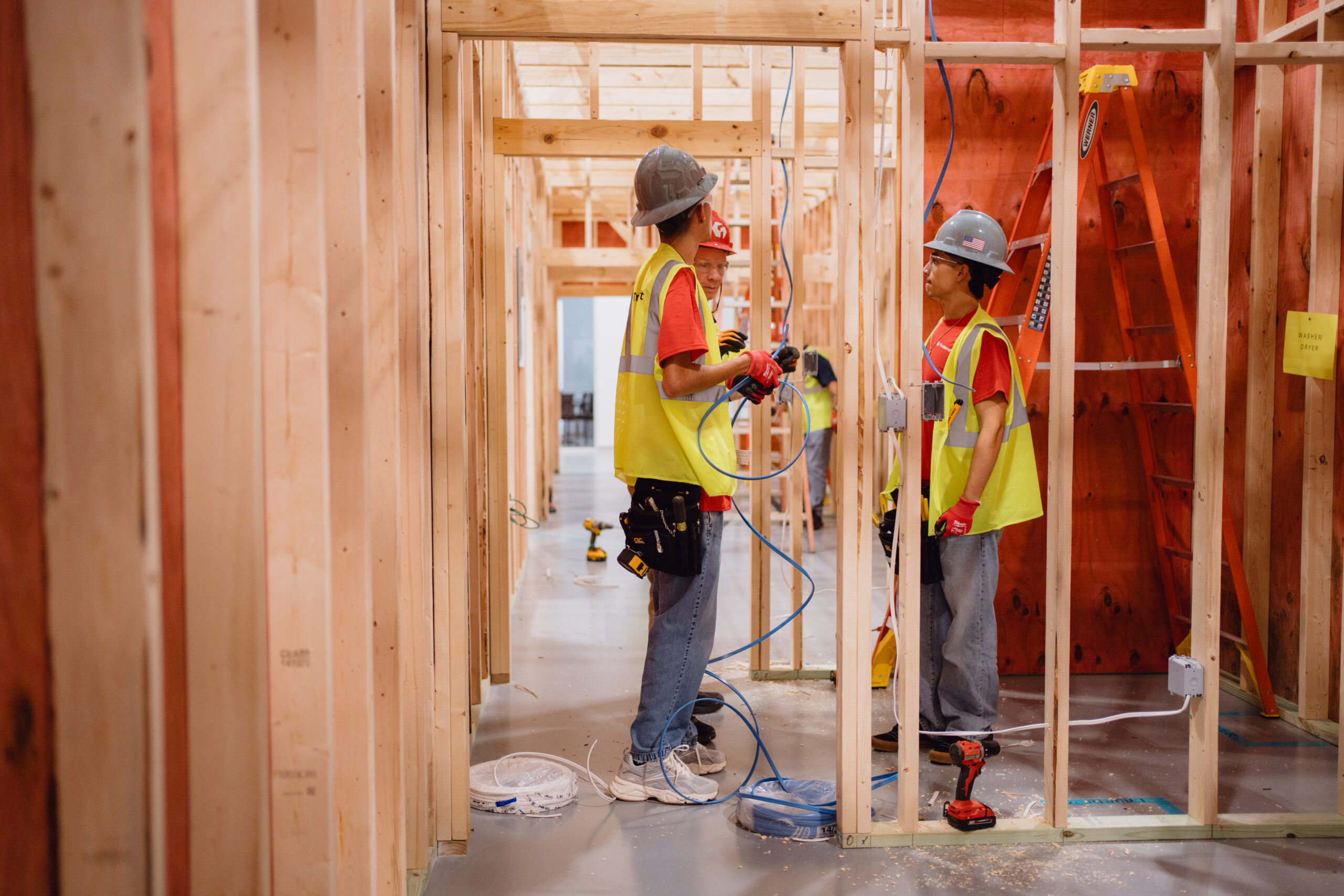 Two Tradecraft electrical students in gray hard hats running electrical wiring through wood studs in a San Antonio lab booth