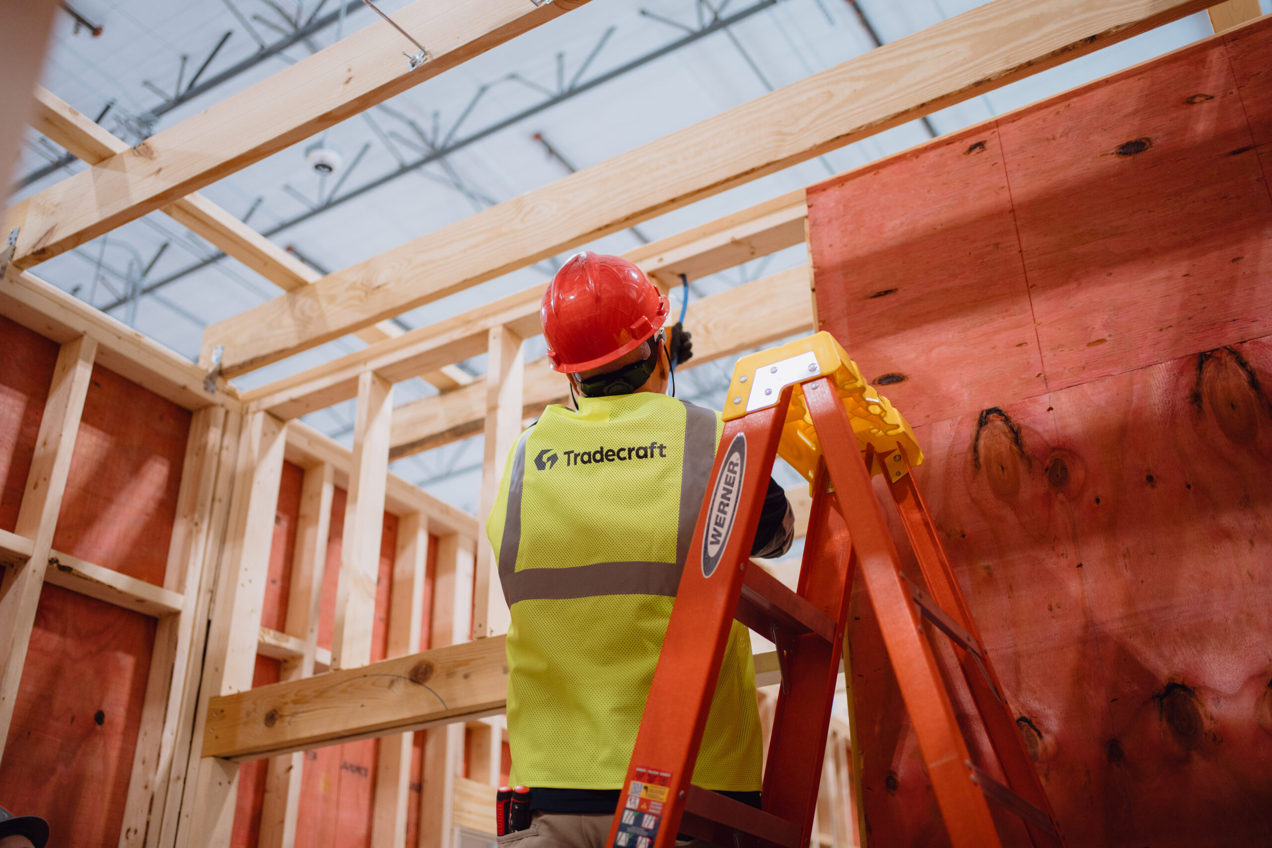 Tradecraft electrical instructor wearing an orange hard hat on a stepladder running electrical wiring through ceiling joists