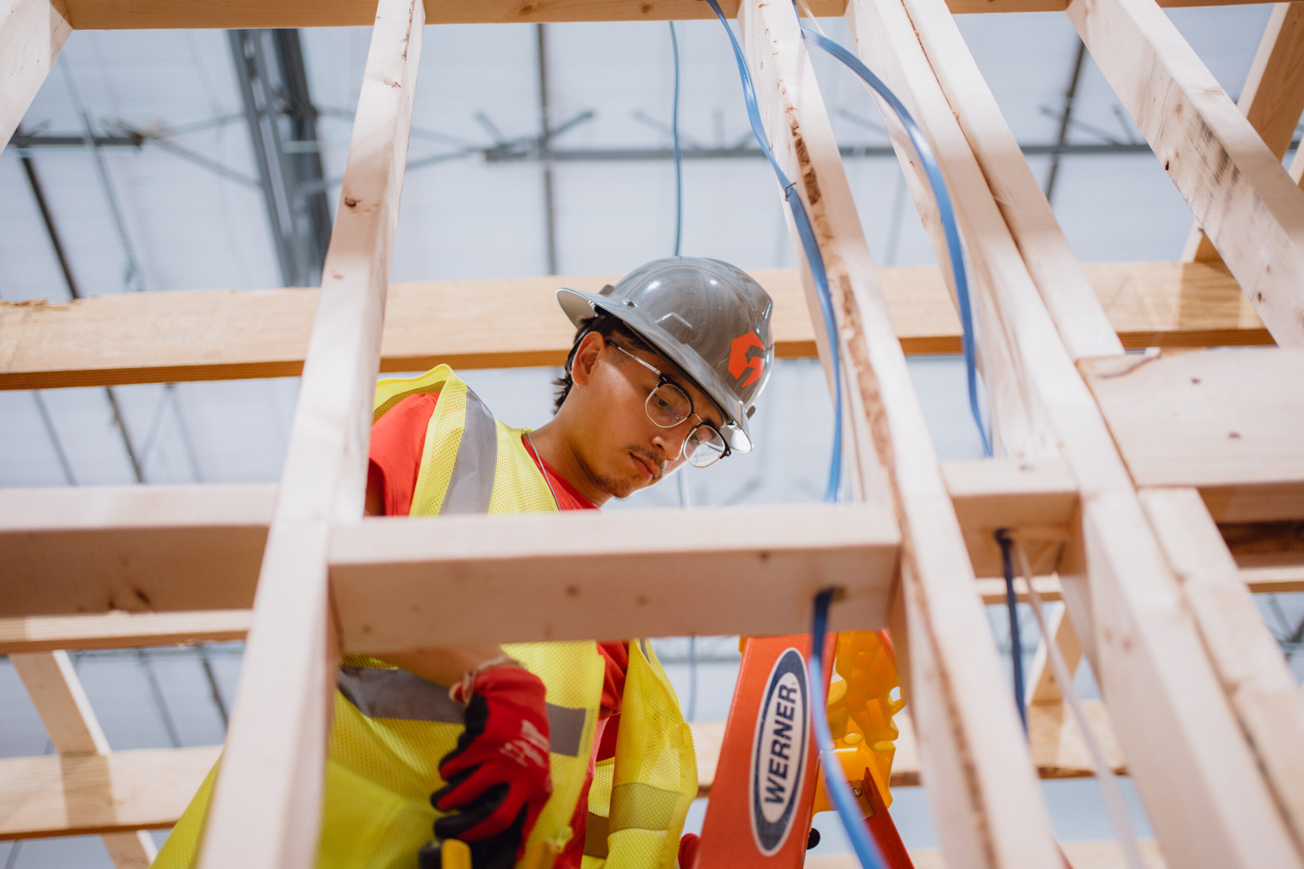 Tradecraft electrical student in a gray hard hat pulling wire through wooden wall framing on a ladder