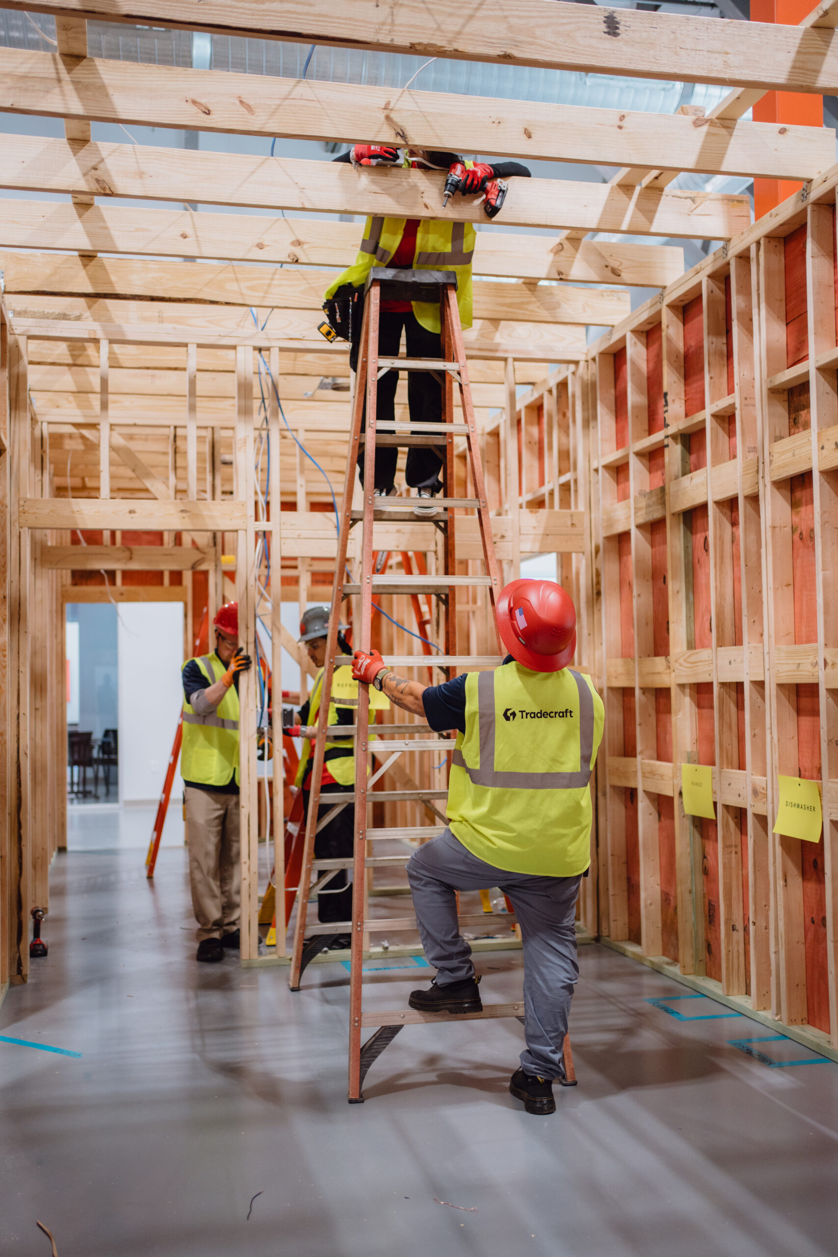 Tradecraft electrical instructor in an orange hard hat guiding students working on ladders in a framed lab booth