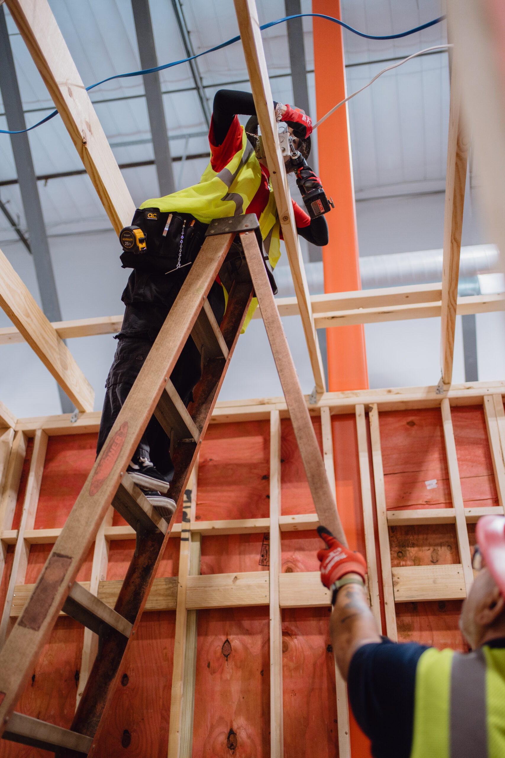 Tradecraft instructor assisting an electrical student on a ladder drilling an electrical junction box into ceiling joists